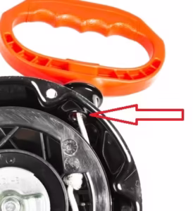 Widok rozrusznika kosiarki od spodu, strzałka wskazuje na przelotkę sznurka szarpaka.