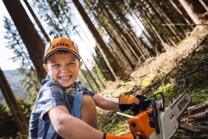Chłopiec w czapce i rękawicach STIHL bawi się w lesie pilarką zabawką STIHL MS 500i.