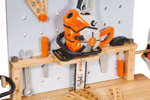 Hełm zabawkowy i rękawice robocze KIDS na dolnej półce drewnianego stołu warsztatowego STIHL.