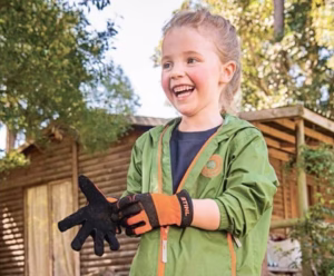 Uśmiechnięta dziewczynka zakłada pomarańczowo-czarne rękawiczki robocze STIHL KIDS.